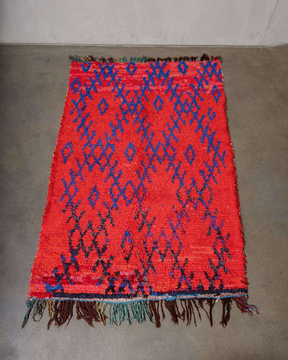 Red and blue patterned rug with fringes on a concrete floor.